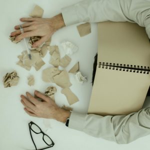 A tired adult man resting his head on a desk cluttered with crumpled papers and an open notebook.